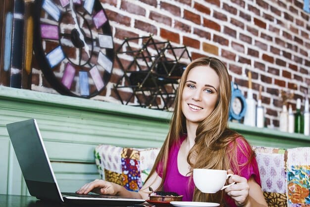 A young woman happily working remotely on her laptop at a vibrant coffee shop, showcasing the flexibility and freedom associated with the creator economy lifestyle. Various creative tools and notebooks are scattered around her, symbolizing her work.
